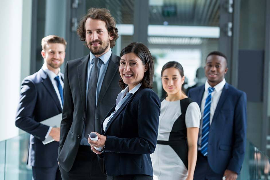 A diverse group of five business professionals in suits stand smiling in a bright modern office corridor, led by a confident woman in a dark blazer holding a document in the foreground.