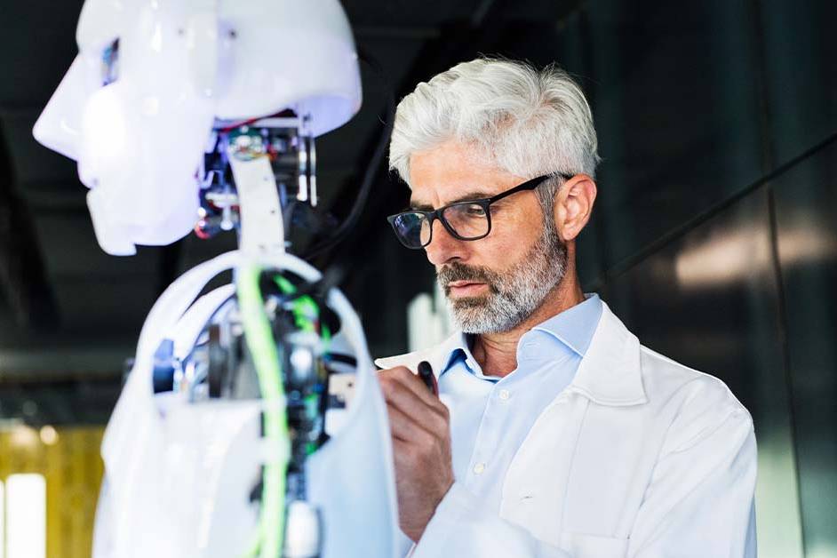 A focused middle-aged male scientist in a white coat and glasses works on a humanoid robot with exposed wiring and circuitry in a modern lab, representing AI and robotics research.