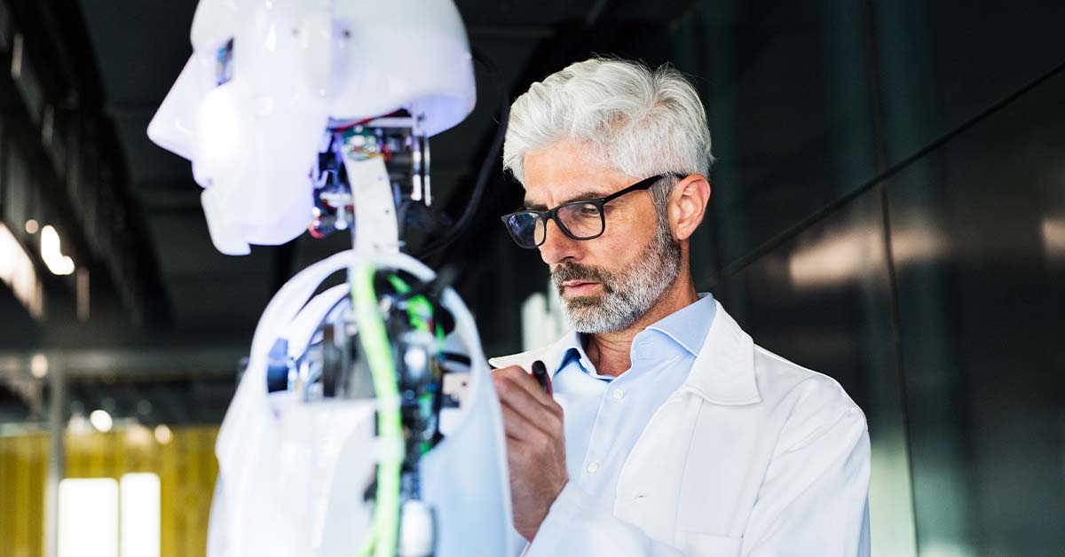 A focused middle-aged male scientist in a white coat and glasses works on a humanoid robot with exposed wiring and circuitry in a modern lab, representing AI and robotics research.
