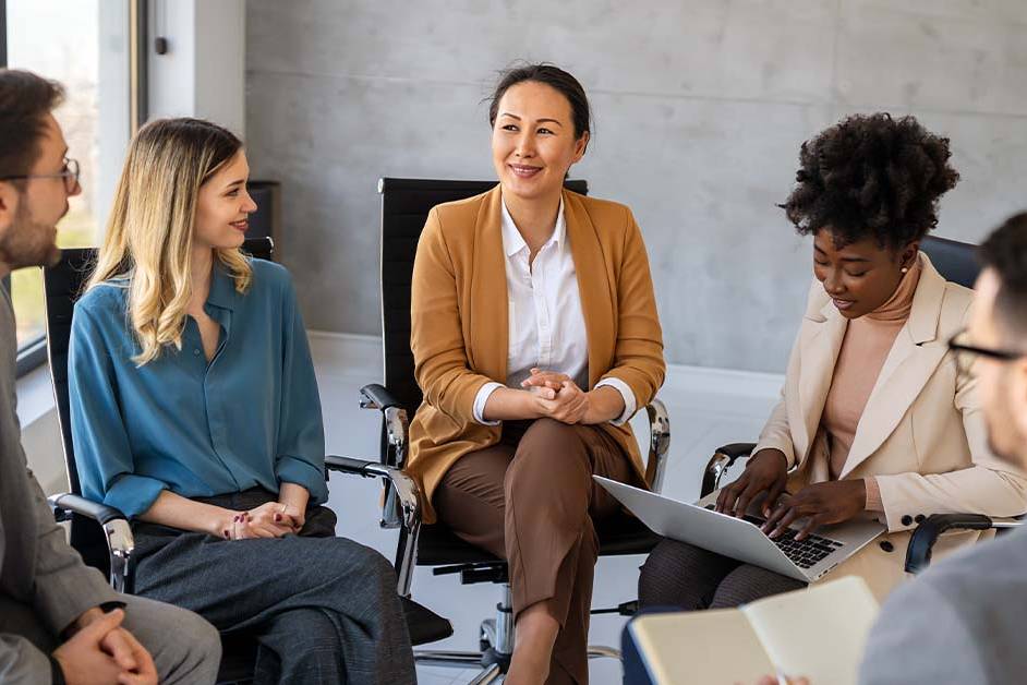 A diverse group of five business professionals sit together in a circle of office chairs, engaged in a warm, collaborative discussion in a modern office.