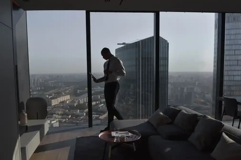 A focused executive standing calmly at a sunlit office window, looking out over a quiet city skyline.