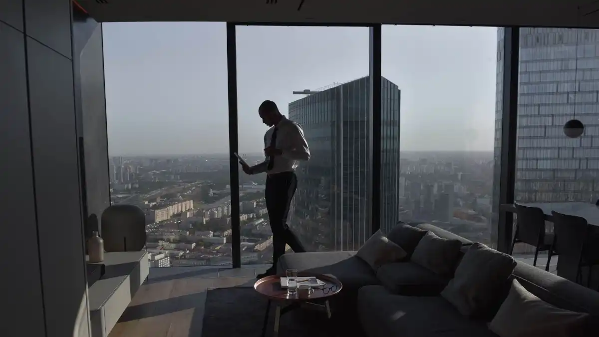 A focused executive standing calmly at a sunlit office window, looking out over a quiet city skyline.