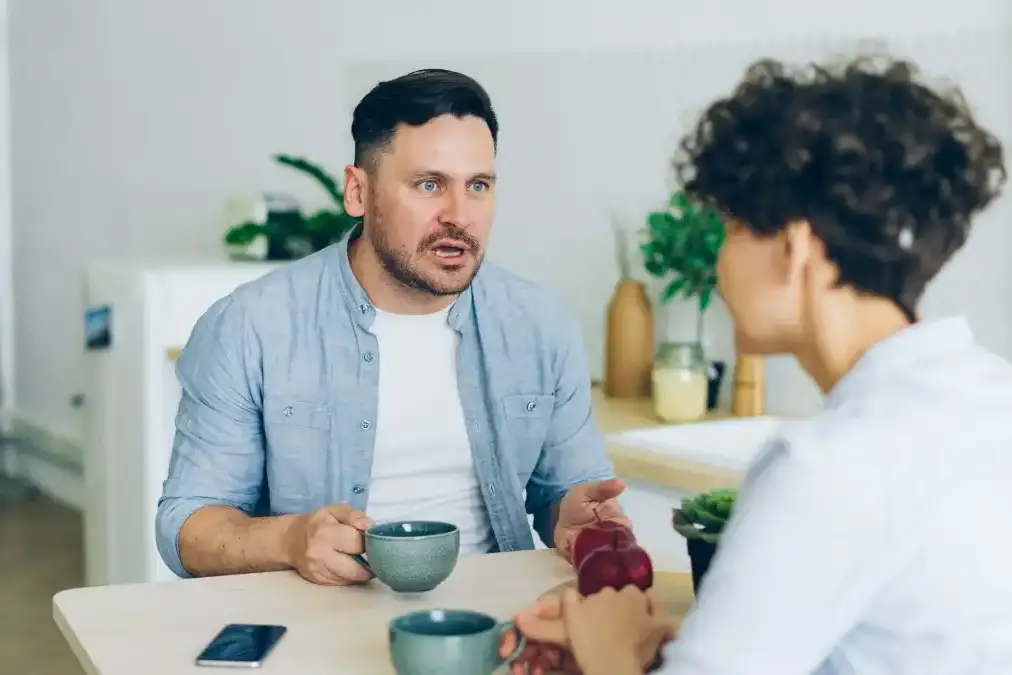A physician couple sitting close together at a kitchen table at night, talking quietly after a long day.
