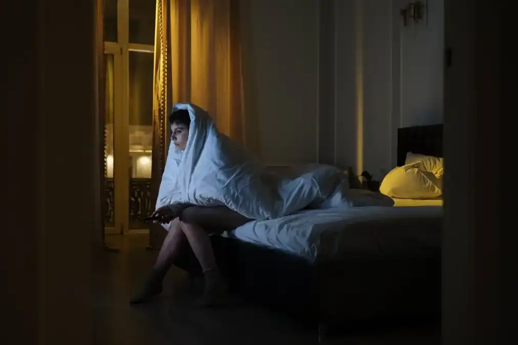 A tired executive sitting awake on the edge of a bed at night, lit only by a dim bedside lamp.