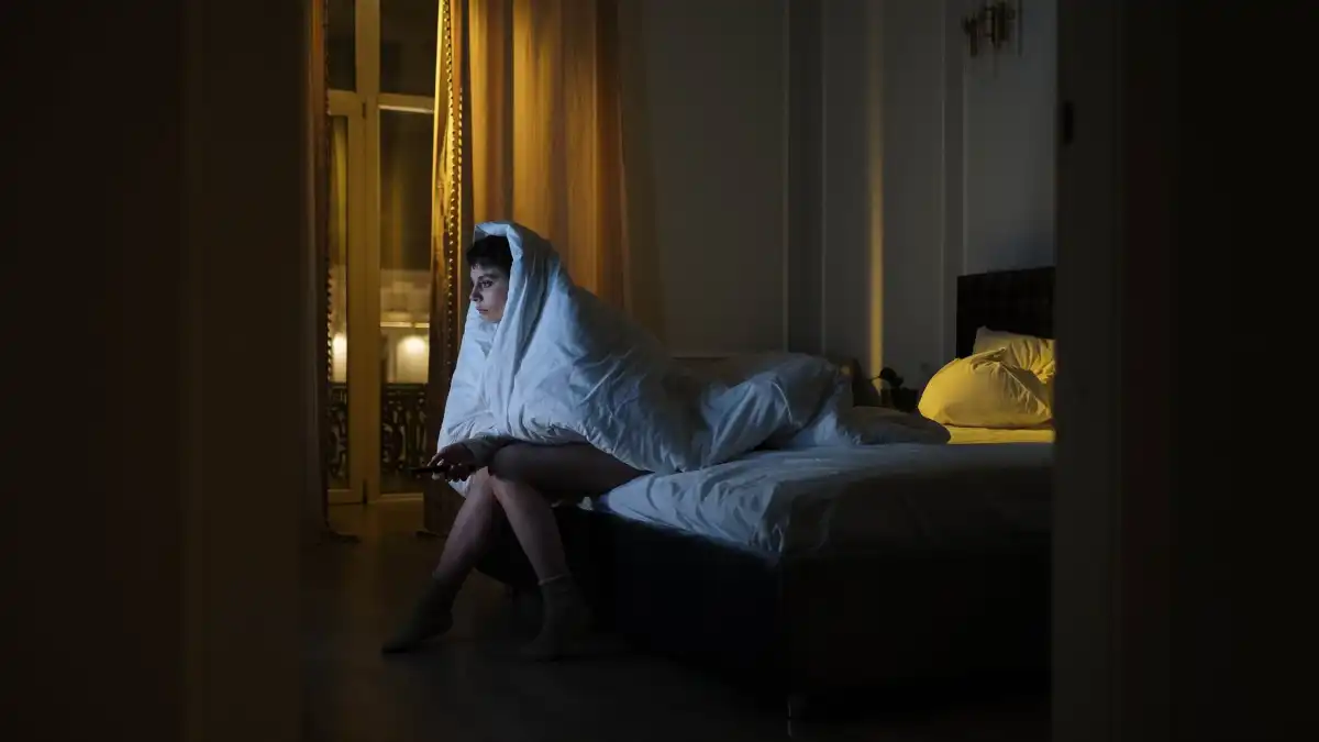 A tired executive sitting awake on the edge of a bed at night, lit only by a dim bedside lamp.