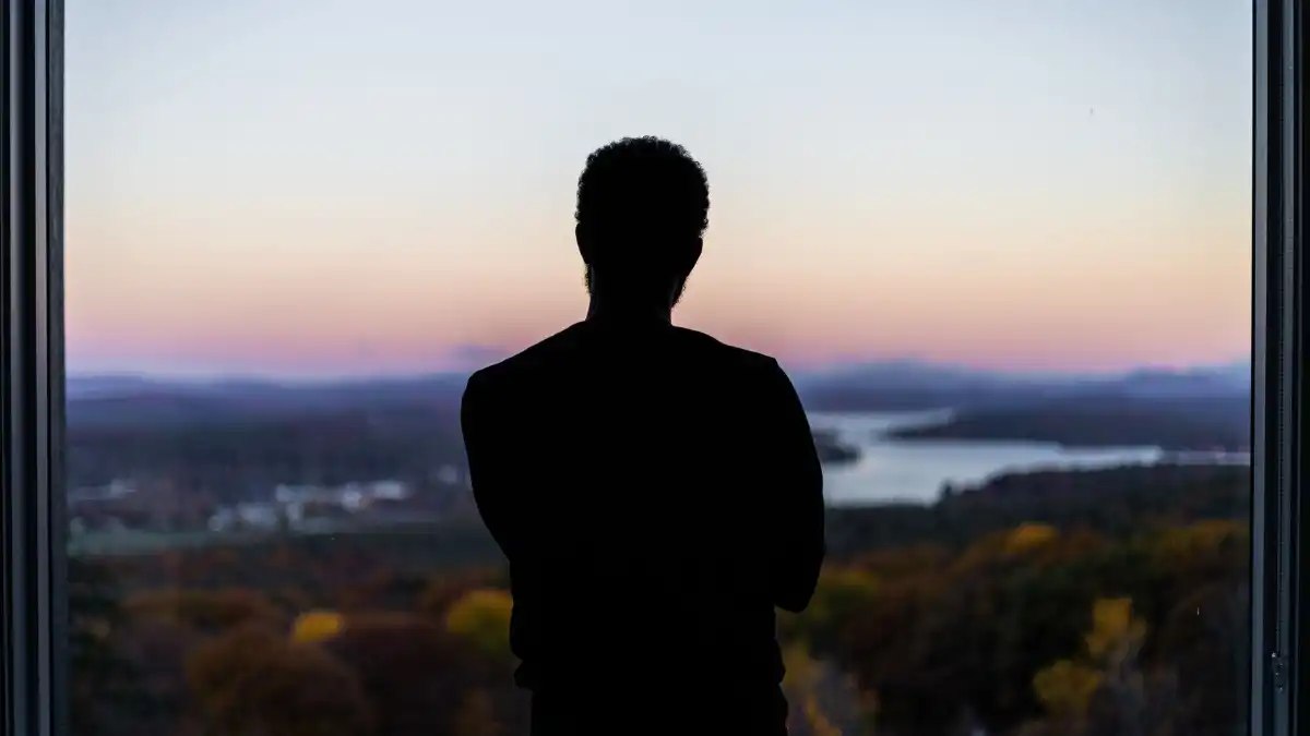 A professional sitting quietly alone by a large window at dusk, reflecting in grief.