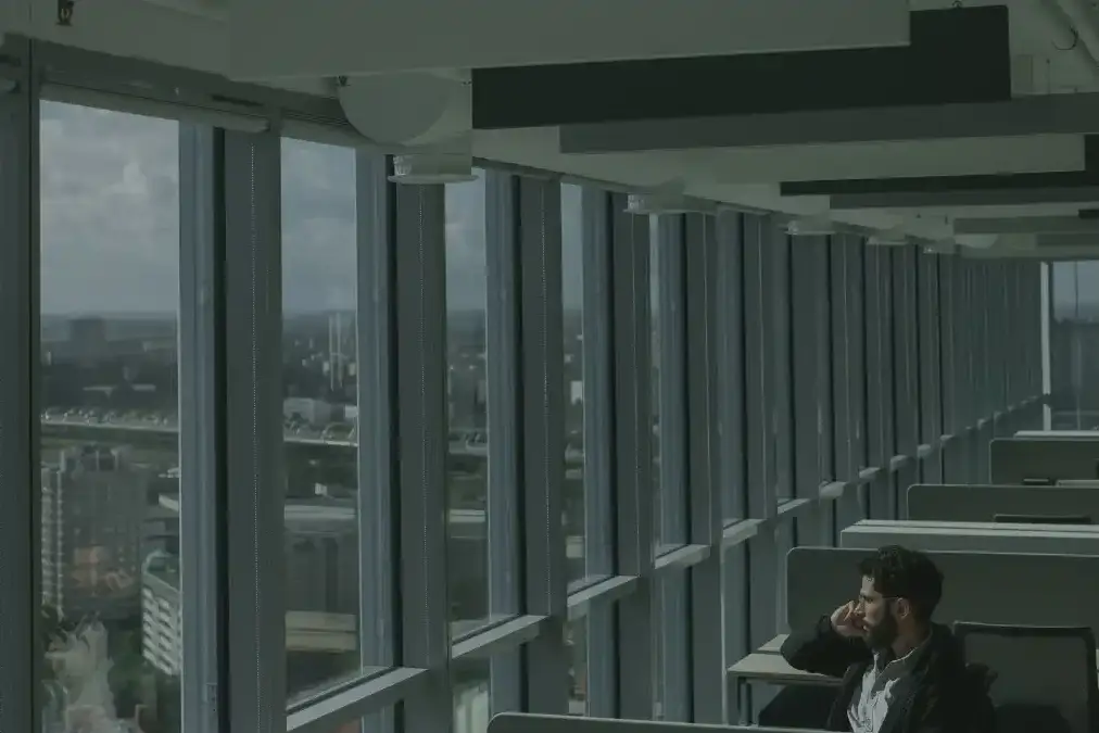 A marketing executive sits alone at a desk by a large office window at dusk, looking thoughtful.