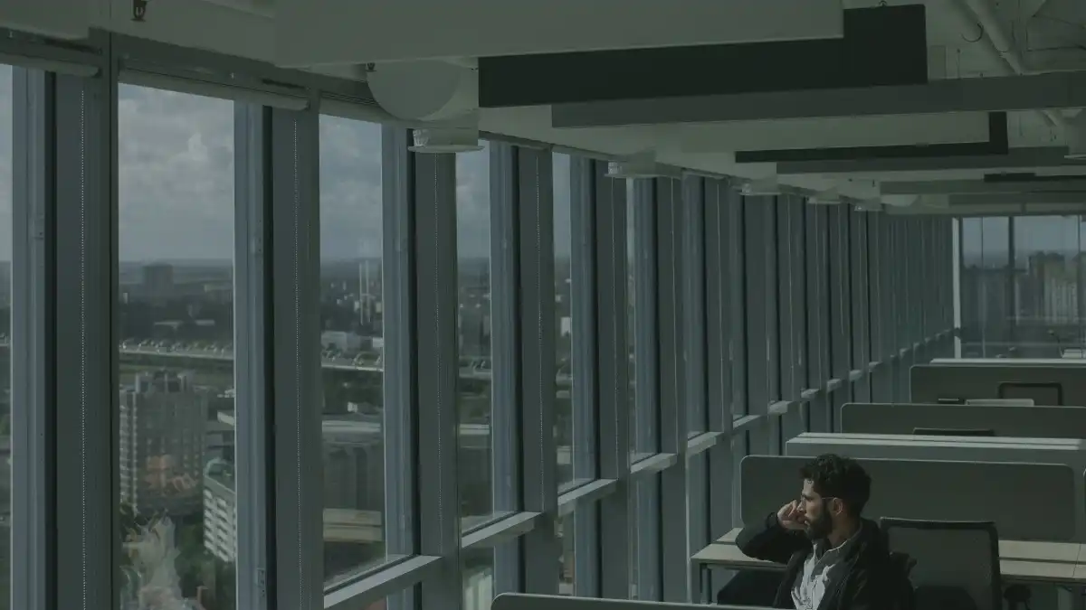 A marketing executive sits alone at a desk by a large office window at dusk, looking thoughtful.
