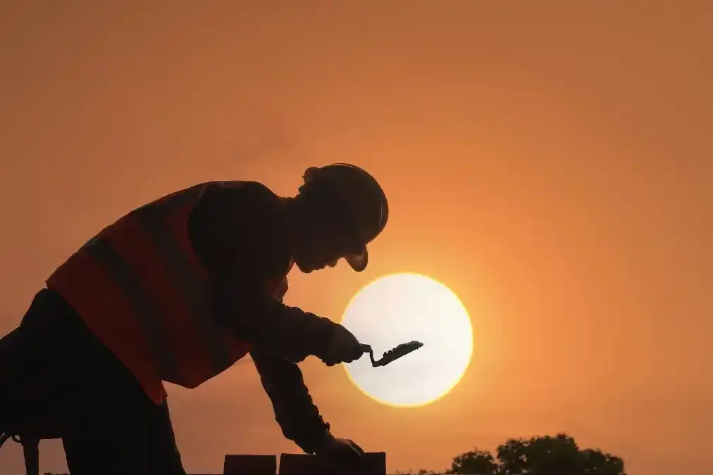 A construction executive in a hard hat looks out over a quiet building site at dawn, deep in thought.
