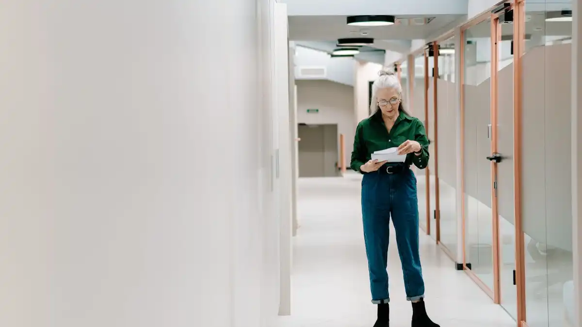 A composed operations executive stands in a quiet corporate hallway, holding a coffee and looking away.