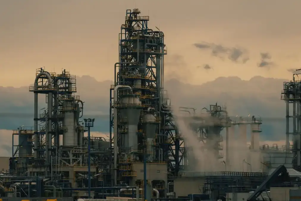 An energy executive stands alone at a window overlooking an industrial skyline at dusk, looking pensive.