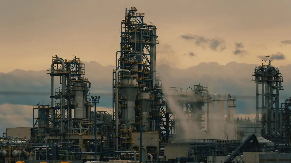 An energy executive stands alone at a window overlooking an industrial skyline at dusk, looking pensive.