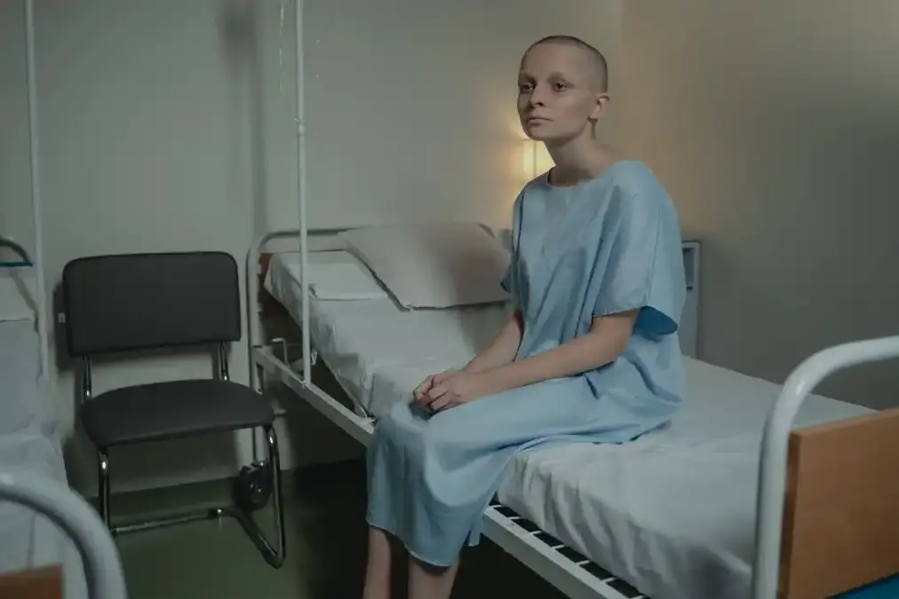 A senior executive sits quietly by a hospital window in daylight, hands clasped, looking out reflectively.