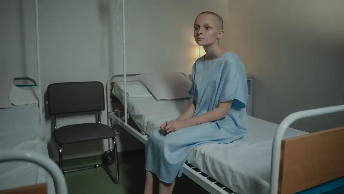 A senior executive sits quietly by a hospital window in daylight, hands clasped, looking out reflectively.
