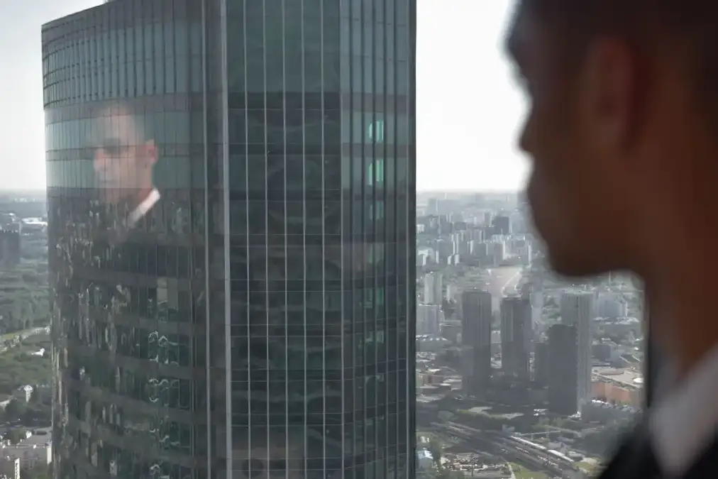 A confident senior leader of color stands alone by a wide office window overlooking a city skyline.