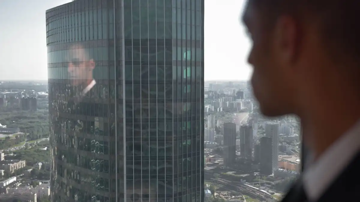 A confident senior leader of color stands alone by a wide office window overlooking a city skyline.