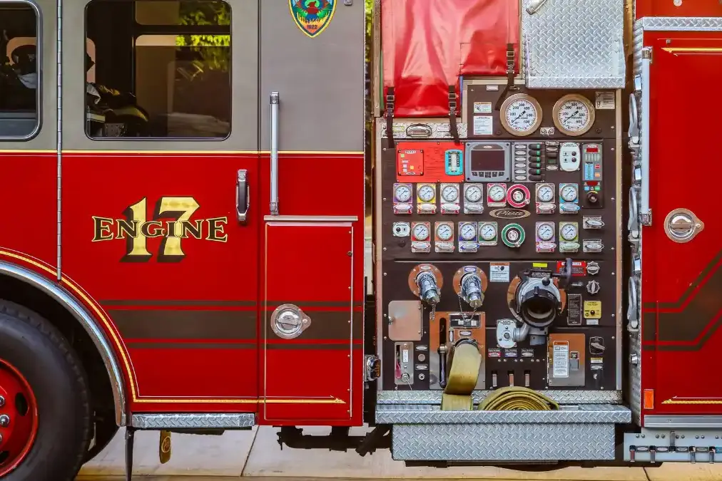 A fire chief stands alone at dusk before a quiet fire station with engines visible behind him.