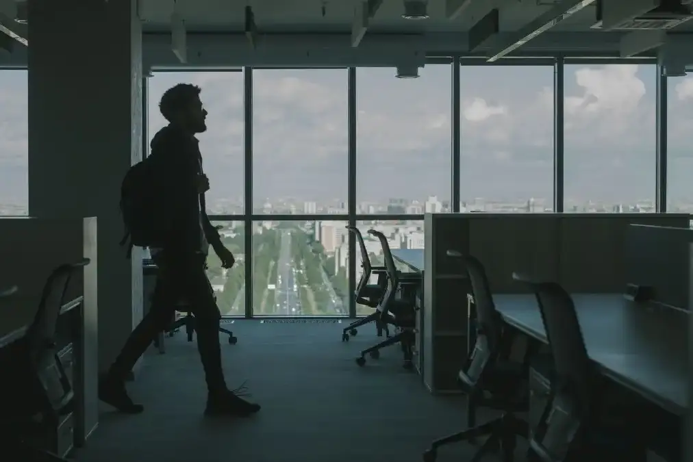 A young professional stands alone in a glass office at dusk looking out over a city skyline.