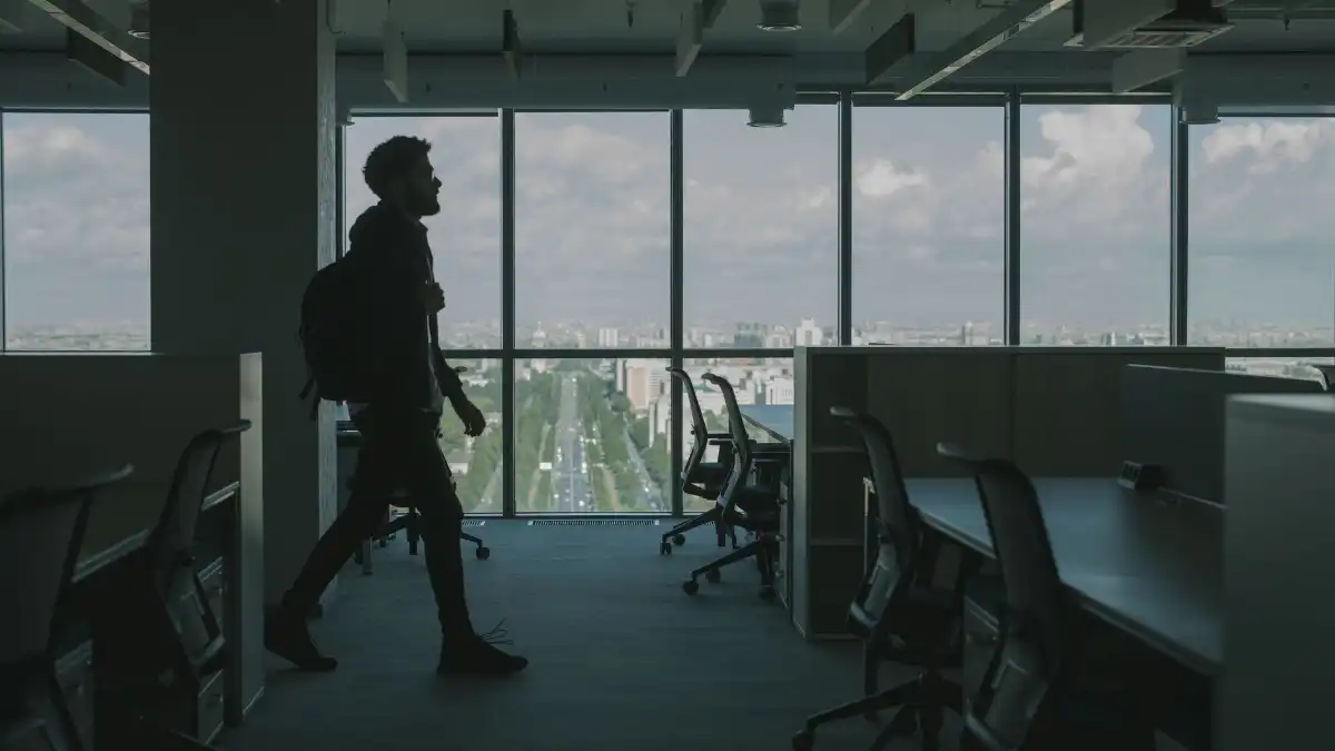 A young professional stands alone in a glass office at dusk looking out over a city skyline.