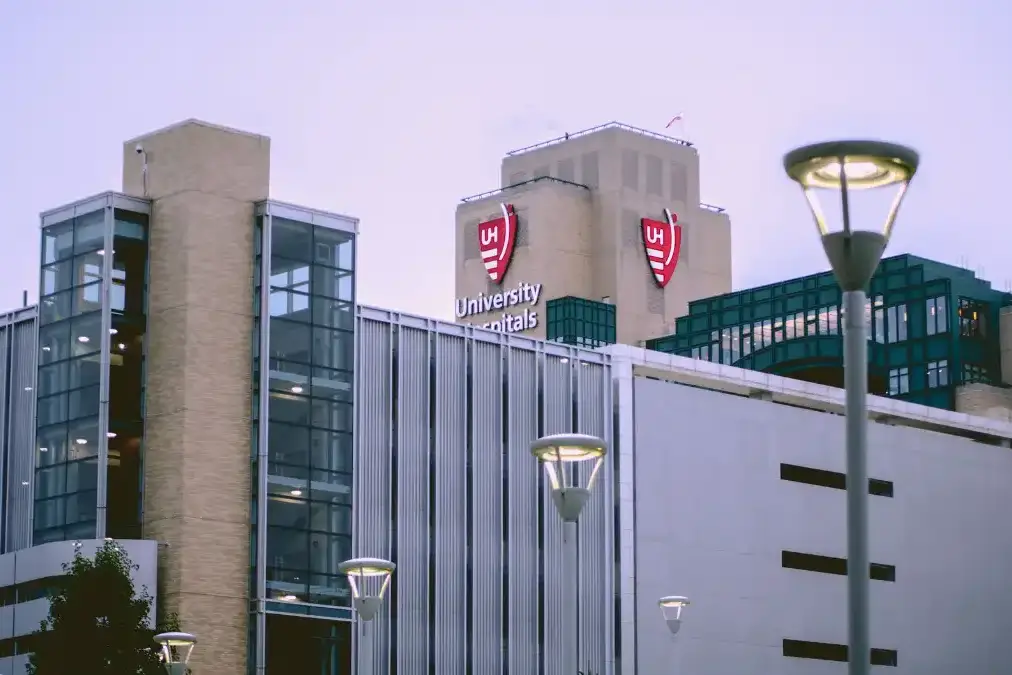 A hospital executive stands alone at a wide window overlooking a medical campus at dusk.