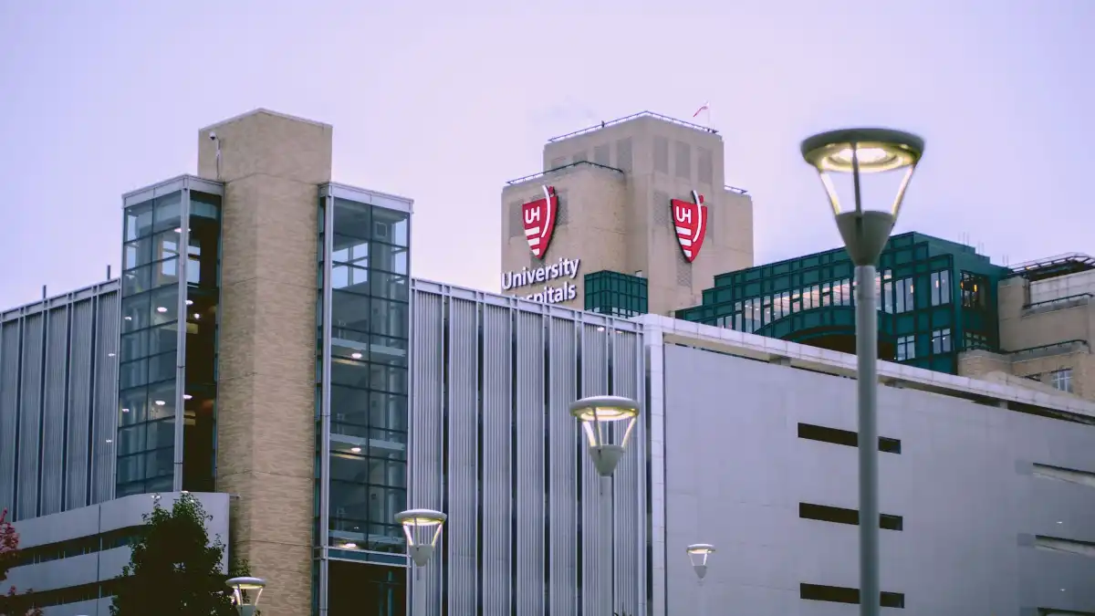 A hospital executive stands alone at a wide window overlooking a medical campus at dusk.