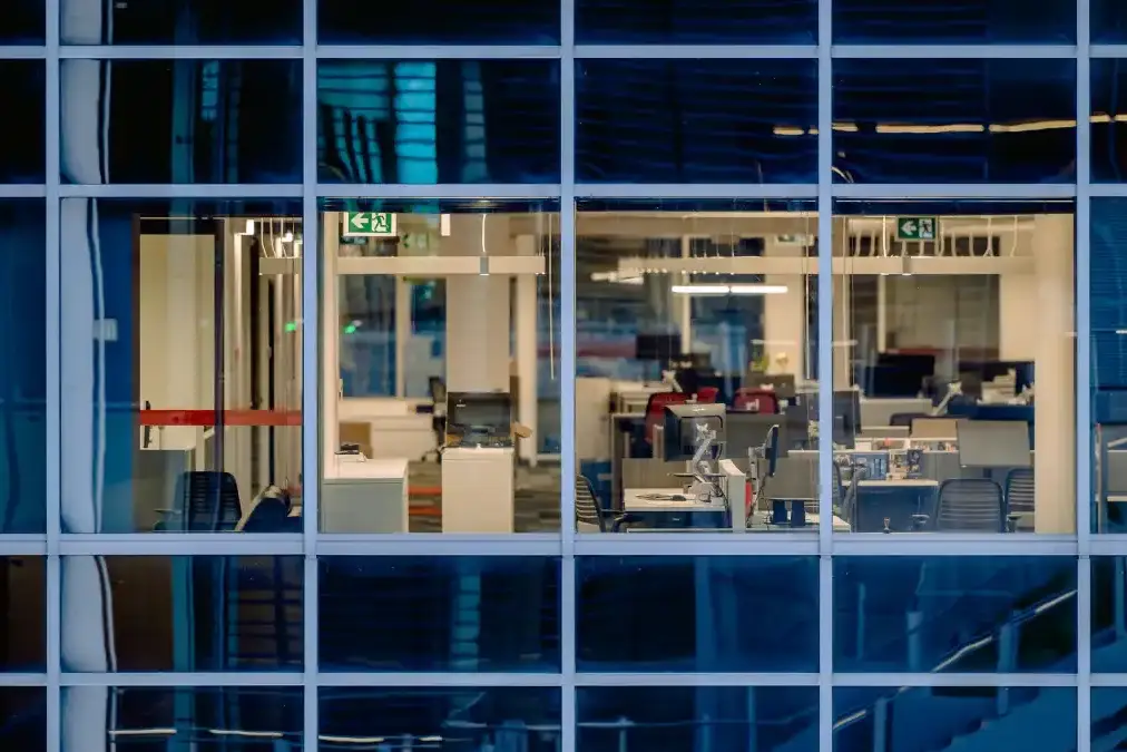An immigrant founder stands alone at a startup office window at dusk looking over a city skyline.