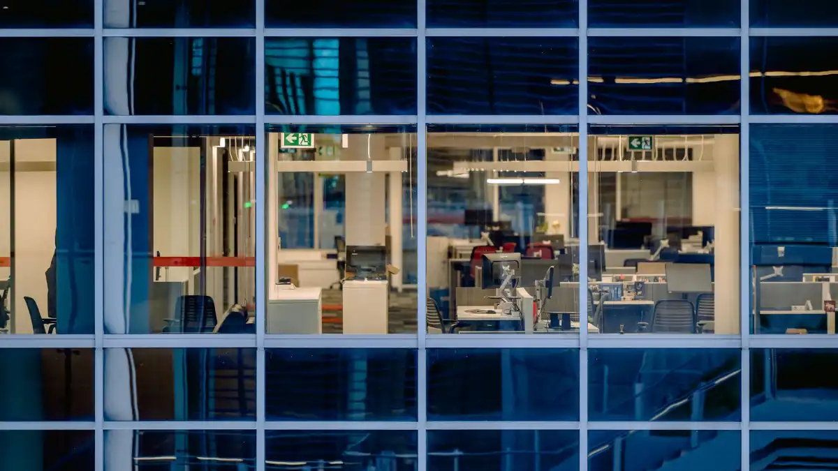 An immigrant founder stands alone at a startup office window at dusk looking over a city skyline.
