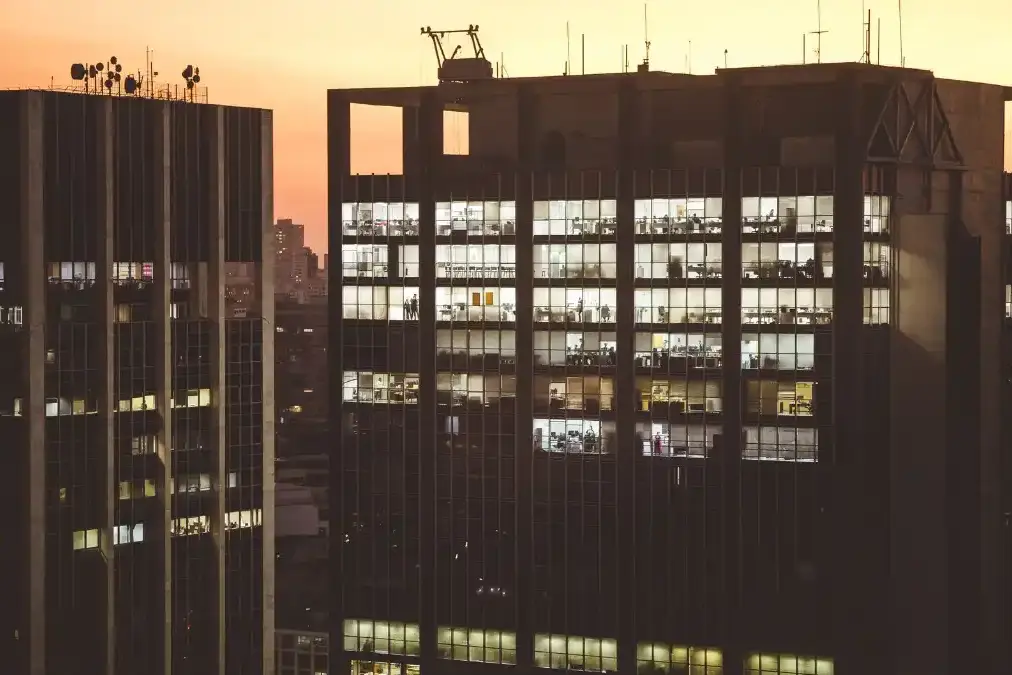 An insurance executive stands alone at a high office window overlooking a financial district at dusk.