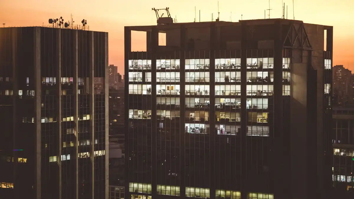 An insurance executive stands alone at a high office window overlooking a financial district at dusk.