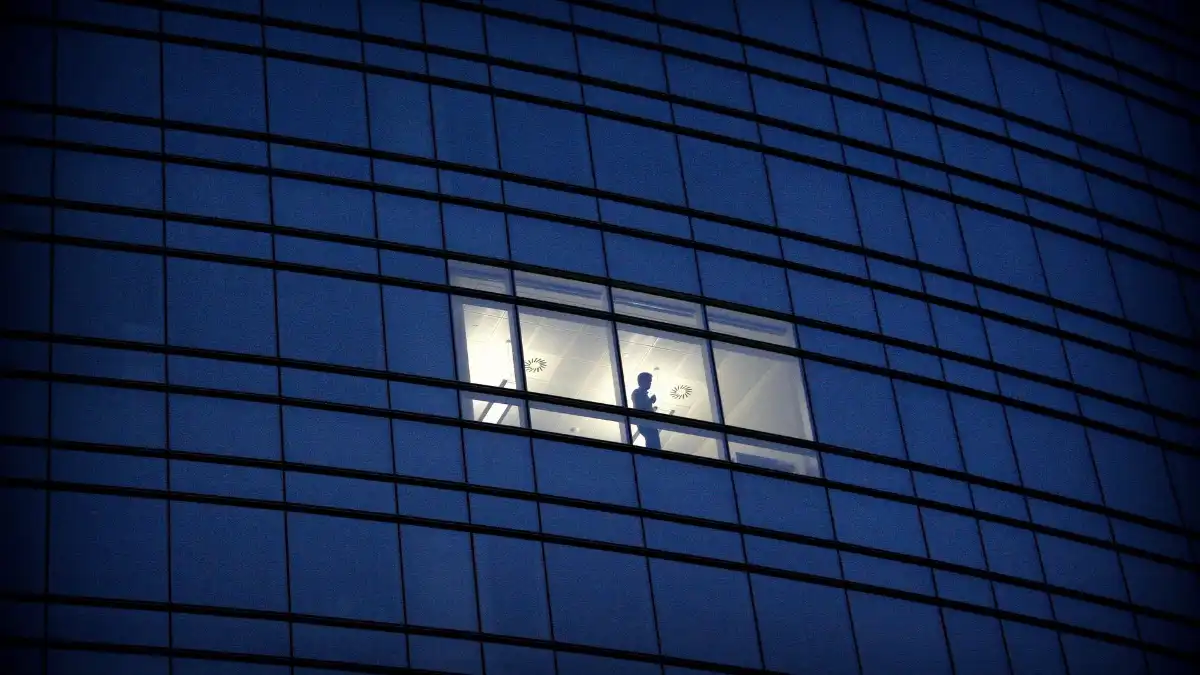Exhausted investment banker alone at a desk by a city office window late at night.