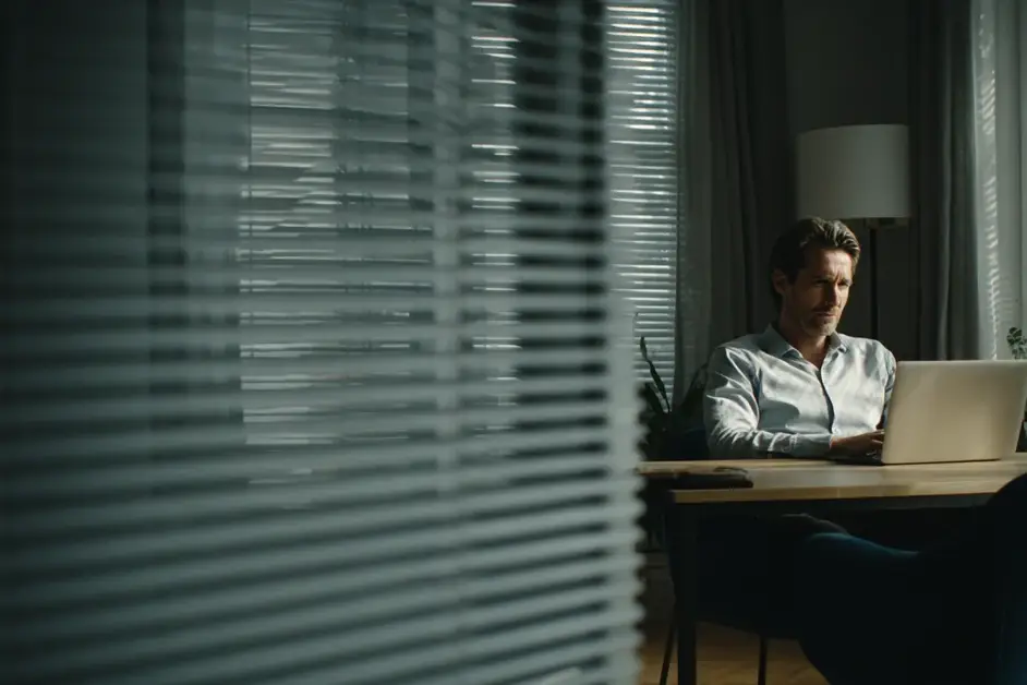 Man in a shirt sitting at a desk in a private office with the blinds half drawn and a laptop open in front of him