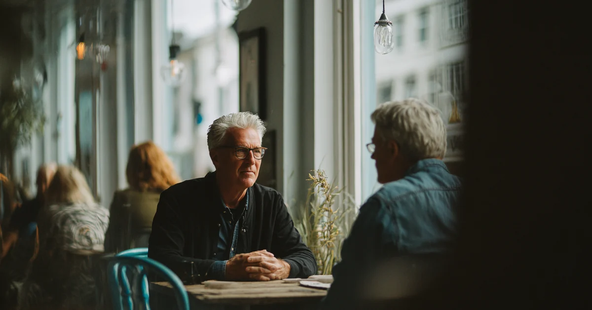 Two people seated across a cafe table by a window, one talking while the other listens with a neutral expression