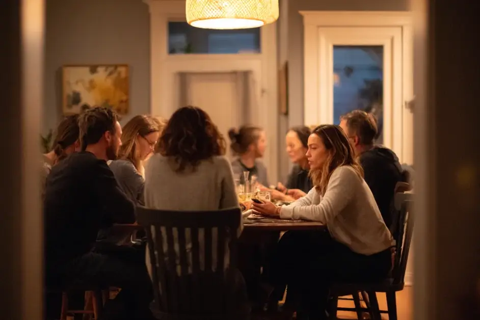 Family gathered around a lit dining table in the evening, one woman at the end holding a phone in her lap and looking away