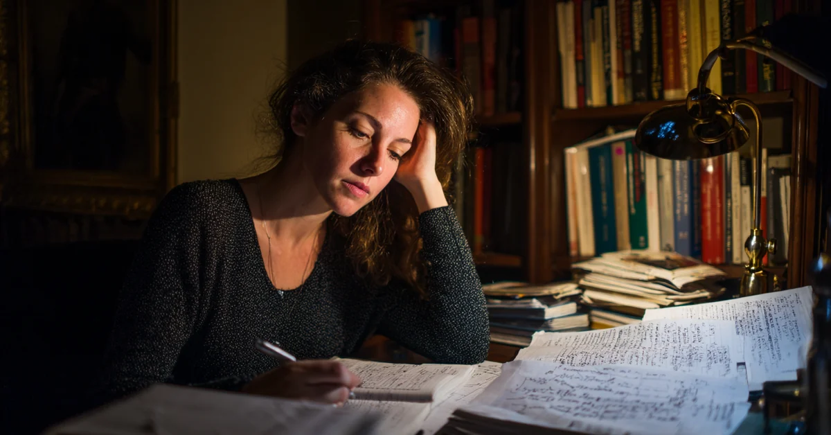 Woman at a desk late at night under one lamp, hand against her temple, surrounded by open books and densely handwritten pages