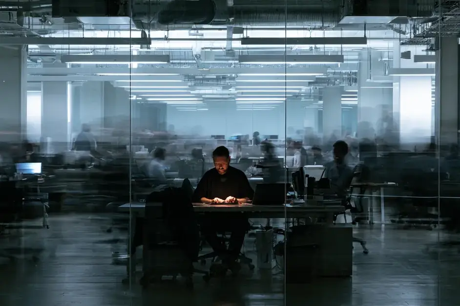 Long-exposure photo of a still, lit worker at a desk surrounded by a blurred, bustling office behind glass