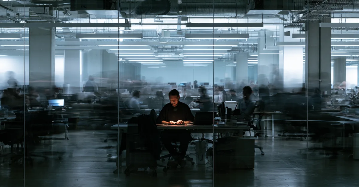 Long-exposure photo of a still, lit worker at a desk surrounded by a blurred, bustling office behind glass