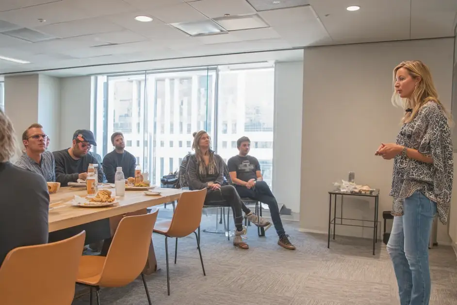 Woman standing to speak at a lunchtime office session while colleagues sit along a table, several chairs empty