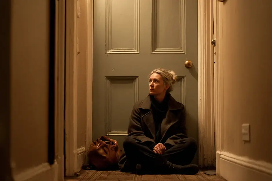 Woman sitting on the floor of a hallway with her coat still on and a bag beside her, back to the closed front door