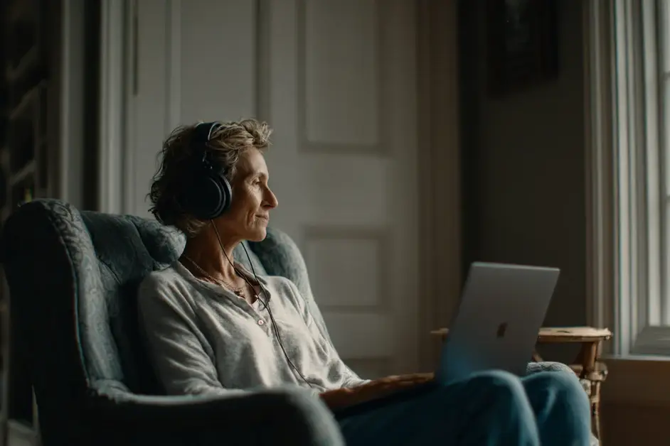 Woman wearing headphones sitting in an armchair at home facing a laptop on a side table