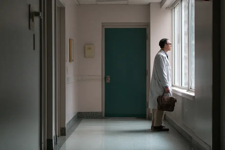 Physician in a white coat standing at a corridor window with a bag in hand, an empty hospital corridor behind