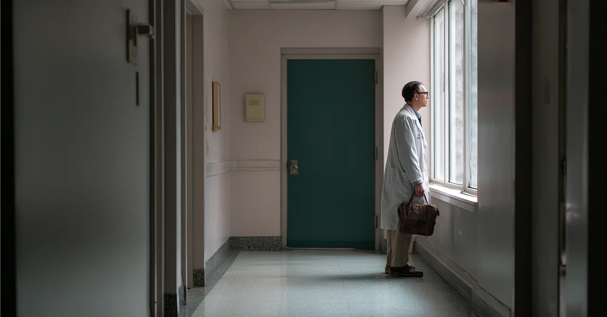 Physician in a white coat standing at a corridor window with a bag in hand, an empty hospital corridor behind