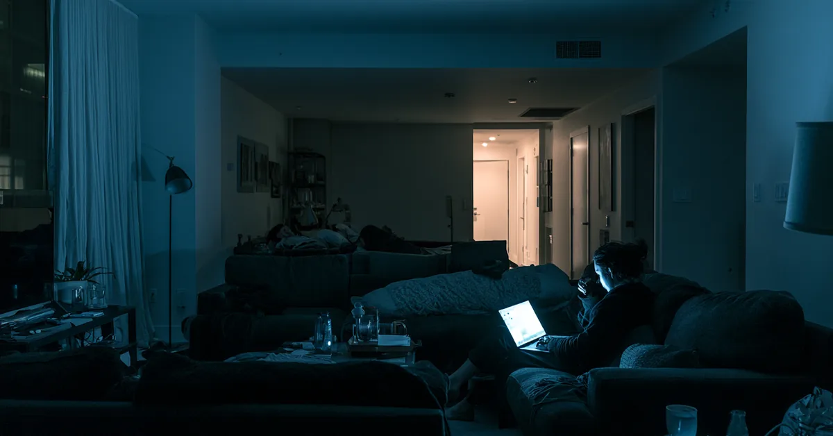 Person on a couch lit only by a laptop screen in a dark apartment living room at 1:20 AM