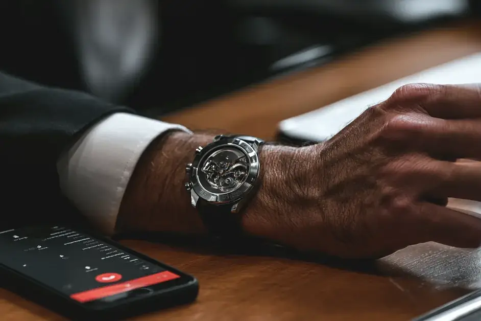 Close-up of a wrist wearing a mechanical watch reaching toward a laptop beside a ringing phone