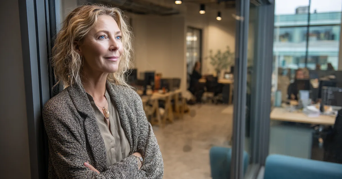 Woman standing at an internal office window looking out over a small working floor with people at desks