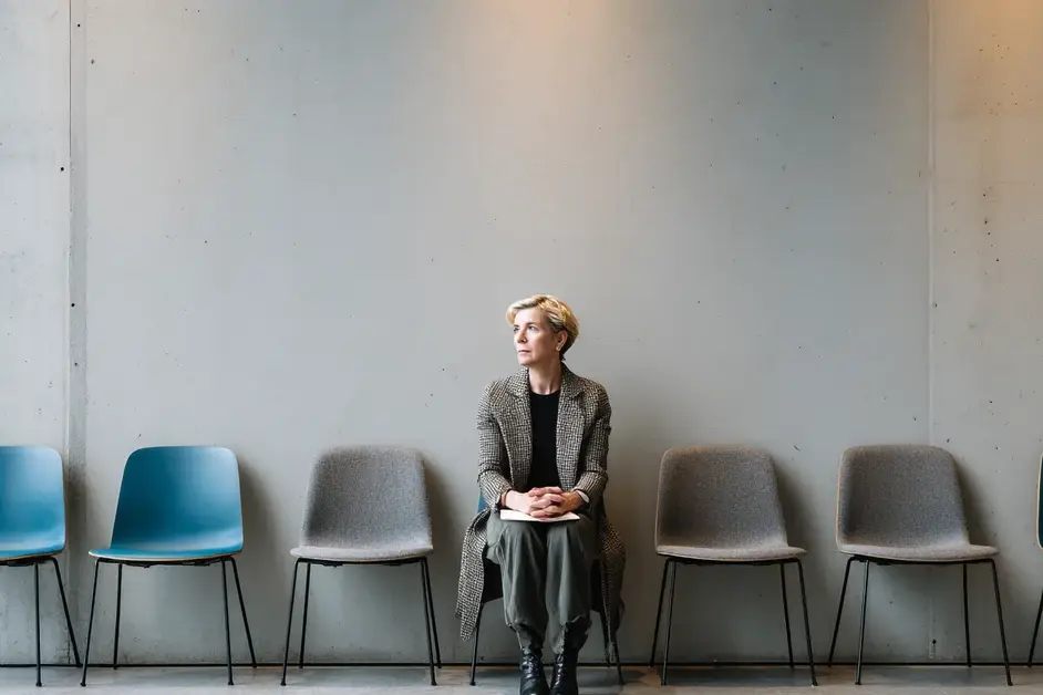 Woman in professional clothing sitting alone in a row of empty waiting-area chairs against a bare concrete wall, hands clasped in her lap