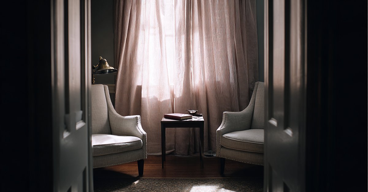 A quiet consulting room seen through a doorway: two linen armchairs facing each other, a small table with a closed notebook, soft light through curtains.