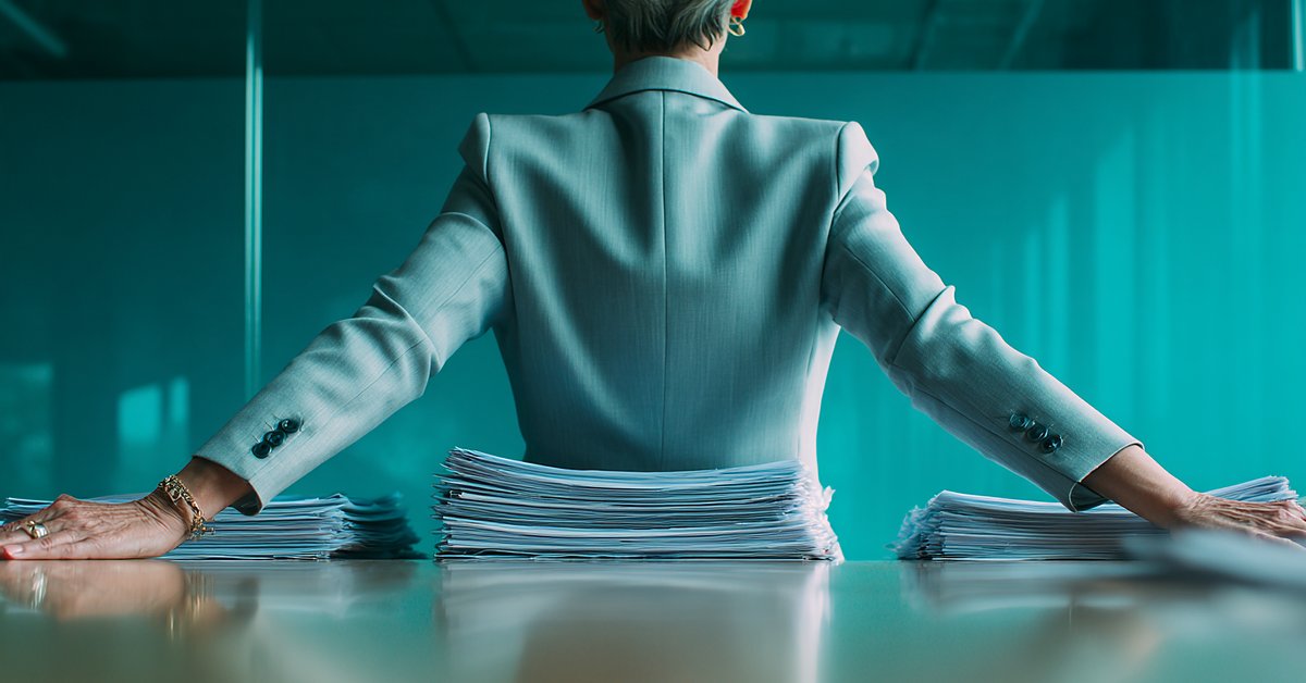 A woman executive seen from behind at a teal conference table, arms braced wide on either side of a tall stack of papers.