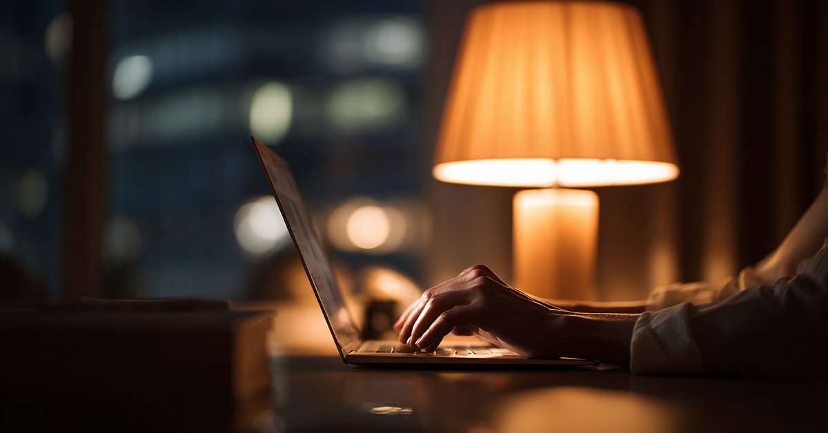 Hand resting on a laptop keyboard at a lamp-lit desk at night, the pause before booking a first therapy session.