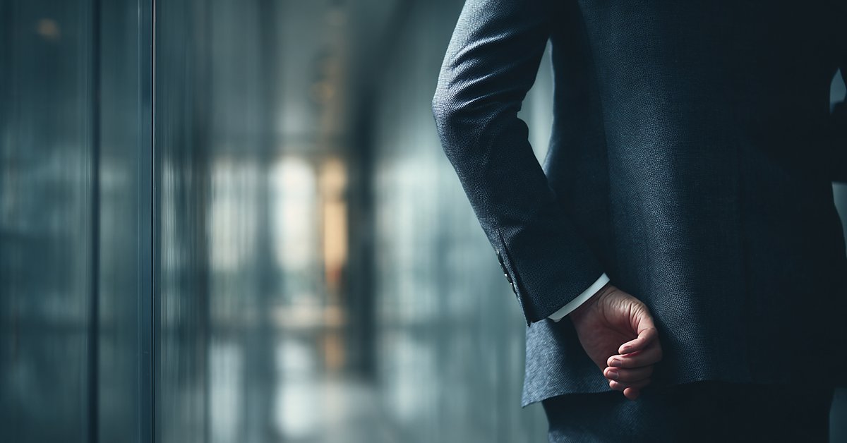 Executive in a dark suit waiting alone in a glass corridor outside a boardroom, hands clasped behind the back before a high-stakes meeting.