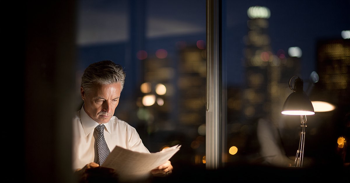 Law firm partner reading a brief under a desk lamp with the Los Angeles skyline behind him at night